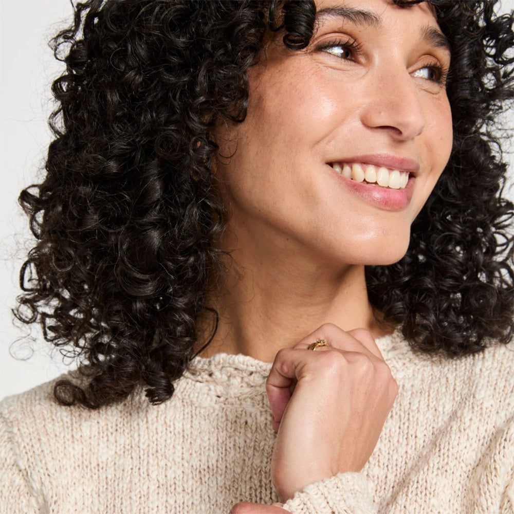 A close-up portrait of a smiling woman with voluminous, dark brown to black curly hair, wearing a cream-colored knit sweater against a plain light background.