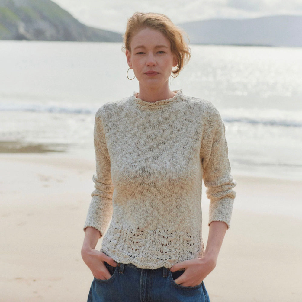 A woman standing on a sandy beach wearing a textured cream-colored knit sweater and blue jeans, with the ocean and distant hills in the background.
