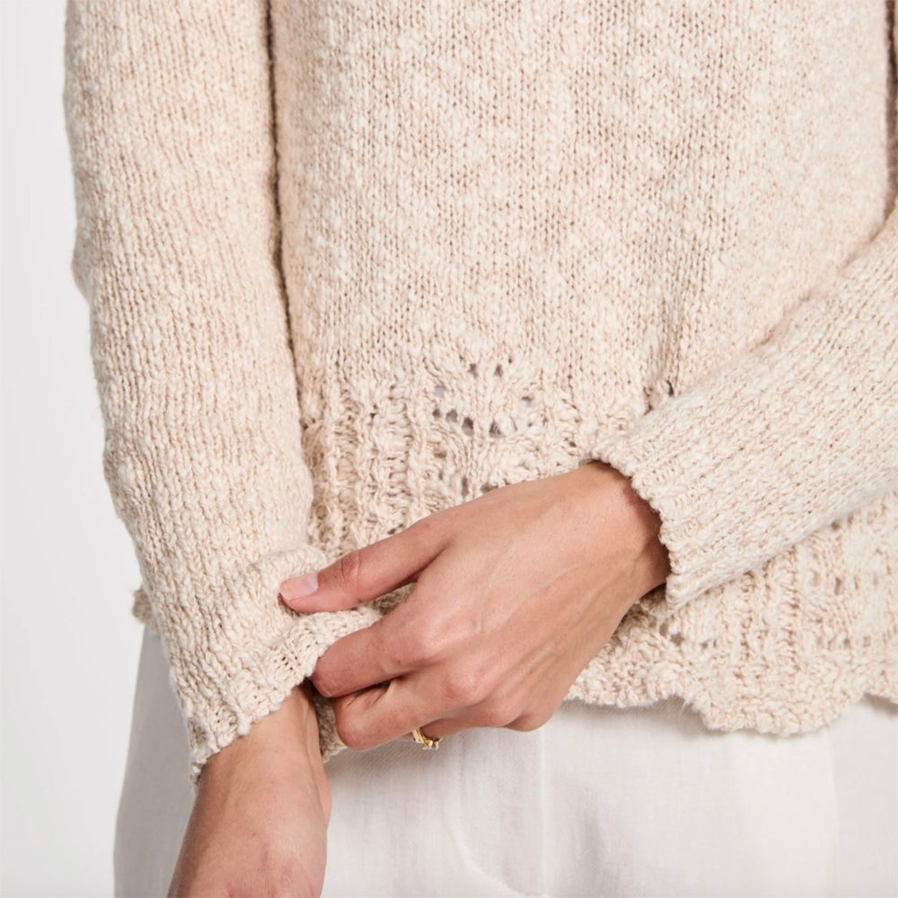 Close-up of a person wearing a chunky knit sweater in a warm cream or oatmeal beige color, with a textured lace-like hem and ribbed cuffs, paired with white pants.