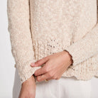 Close-up of a person wearing a chunky knit sweater in a warm cream or oatmeal beige color, with a textured lace-like hem and ribbed cuffs, paired with white pants.