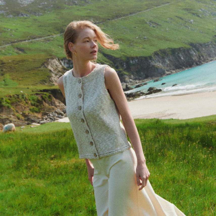 A woman stands in a grassy coastal landscape wearing a sleeveless textured light beige vest with large buttons down the front and cream wide-leg trousers. The background features green hills, a sandy beach, and turquoise water.