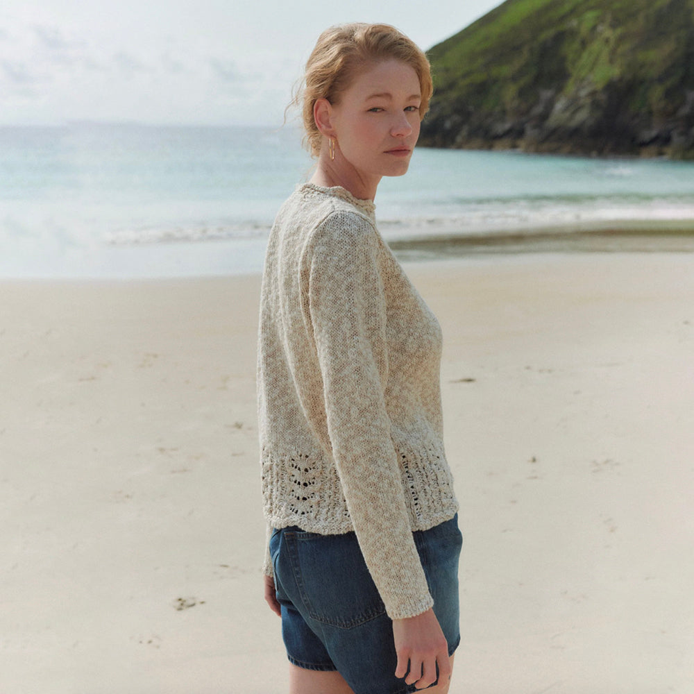 A woman standing on a sandy beach wearing a textured cream-colored knit sweater and denim shorts, with the ocean and green cliffs in the background.