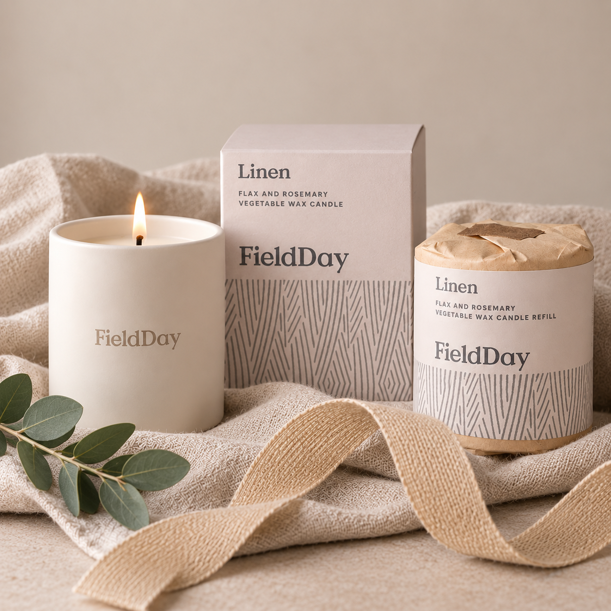 A FieldDay linen scented candle set including a lit white soy wax candle, its matching outer box, and a refill jar wrapped in kraft paper, styled on a beige linen cloth with a ribbon and eucalyptus leaves.