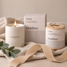 A FieldDay linen scented candle set including a lit white soy wax candle, its matching outer box, and a refill jar wrapped in kraft paper, styled on a beige linen cloth with a ribbon and eucalyptus leaves.