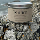 Candle tin labeled Tenfire Coastal Mist on a rock.